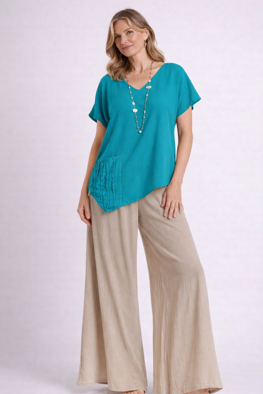A woman poses in the Oh My Gauze Polly Glacier Top T433, a turquoise cotton gauze top with an asymmetrical hem and ruffle detail, styled with beige wide-leg pants and a long beaded necklace against a light background.