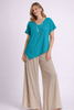 A woman poses in the Oh My Gauze Polly Glacier Top T433, a turquoise cotton gauze top with an asymmetrical hem and ruffle detail, styled with beige wide-leg pants and a long beaded necklace against a light background.