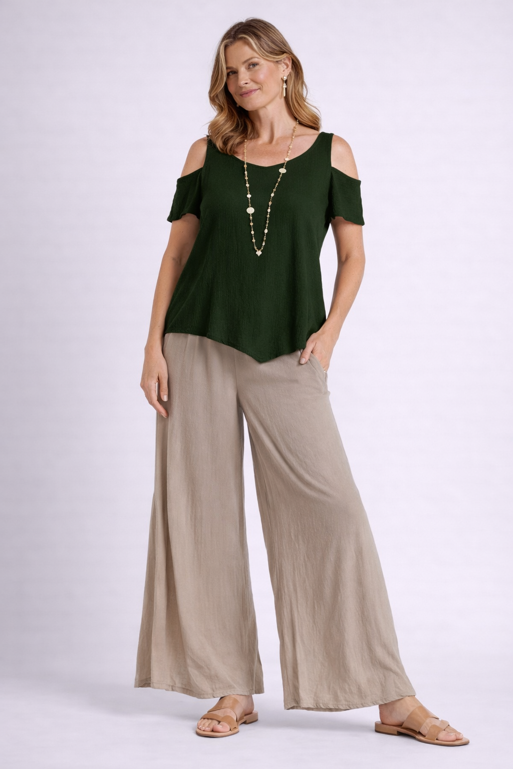 A woman wears the Oh My Gauze Dana Fern Top T150, beige wide-leg pants, tan sandals, and a beaded necklace. She stands against a light background, posing with one hand in her pocket and smiling slightly.