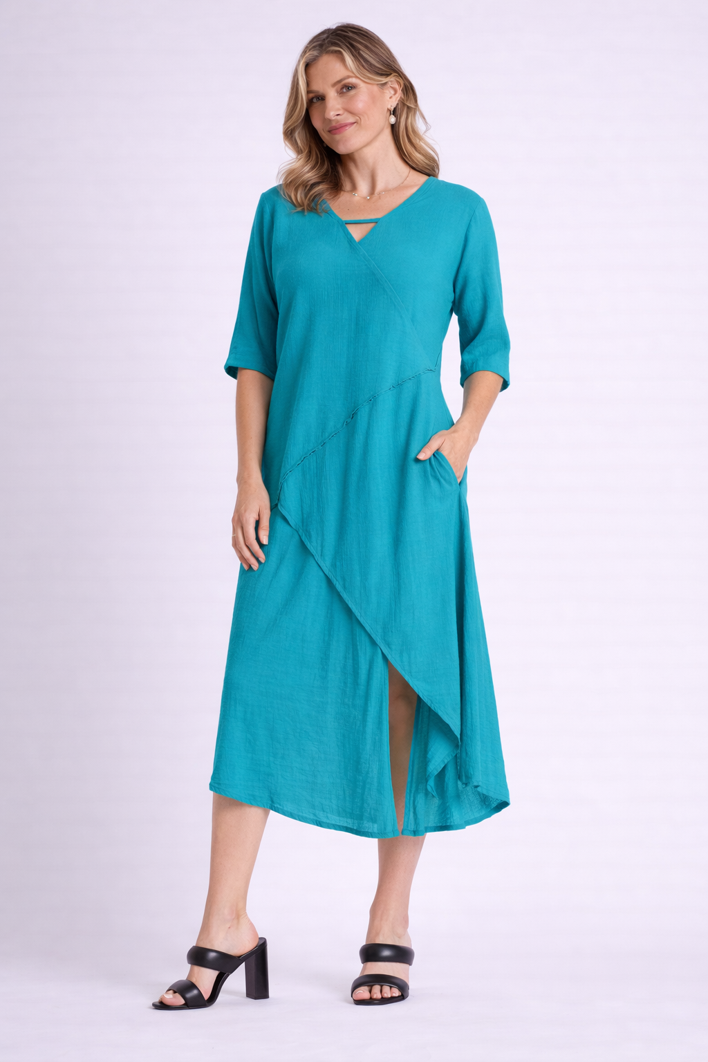 A woman smiles in the Oh My Gauze Dazzle Glacier Dress T706—a teal, knee-length, asymmetrical cotton gauze dress with half sleeves and a front slit—paired with black heeled sandals. One hand rests casually in her pocket.
