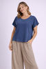 A woman smiles softly against a light purple background, wearing the Oh My Gauze Olga Neptune Top T668—a loose blue cotton blouse with an overlapping design—paired with light beige wide-leg pants for versatile styling.