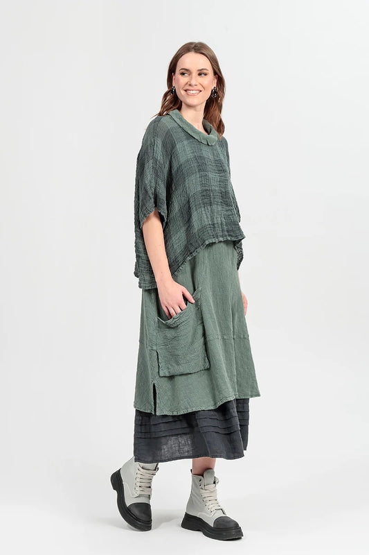 A woman in a BOXY LARGE CHECK SAGE GAUZE LINEN BLOUSE by Paolo Tricot, with a long skirt and light gray lace-up boots, stands smiling against a plain white background, looking to the side.