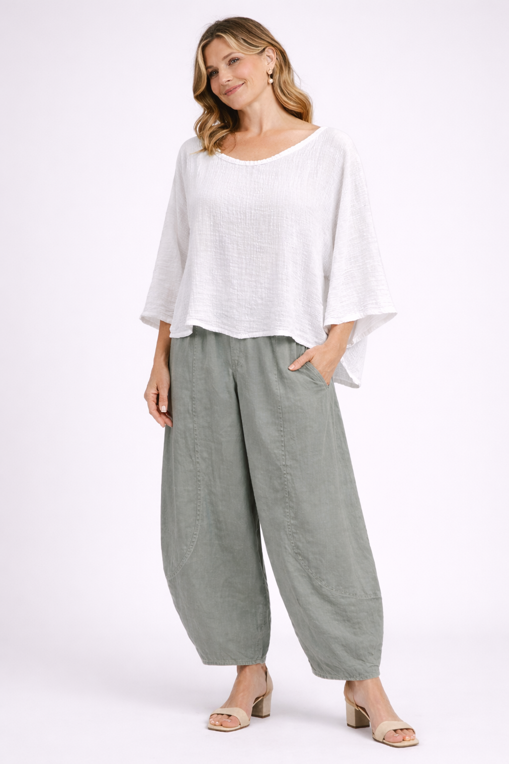 A smiling woman with wavy blonde hair and gold hoop earrings wears Paolo Tricot’s TAPER WIDE COTTON TROUSER - GRIZAS in cream green, paired with a loose white long-sleeve top and beige heels, against a plain light background.