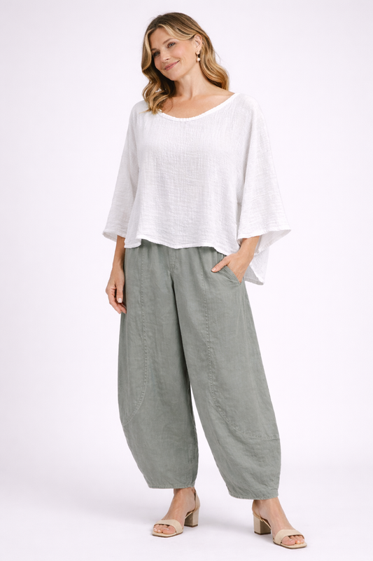 A smiling woman with wavy blonde hair and gold hoop earrings wears Paolo Tricot’s TAPER WIDE COTTON TROUSER - GRIZAS in cream green, paired with a loose white long-sleeve top and beige heels, against a plain light background.