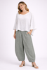 A smiling woman with wavy blonde hair and gold hoop earrings wears Paolo Tricot’s TAPER WIDE COTTON TROUSER - GRIZAS in cream green, paired with a loose white long-sleeve top and beige heels, against a plain light background.