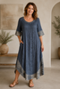 A woman in a blue Dress Mylene 9047 by TaraVao stands indoors on a light floor, smiling with one hand in her pocket. She wears sandals, with potted plants and neutral artwork complementing this versatile TaraVao style.