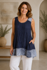 A woman with wavy brown hair smiles indoors, wearing the TaraVao Sleeveless Top Mylene 3084—navy blue with a patterned hem—and white pants. The setting includes potted plants and neutral decor.