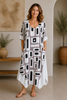A woman stands indoors in the TaraVao Dress Magali 9047, a loose, knee-length Ecovero viscose dress with a geometric black, white, and gray pattern. Barefoot in sandals, she smiles amid potted plants and minimalist decor.