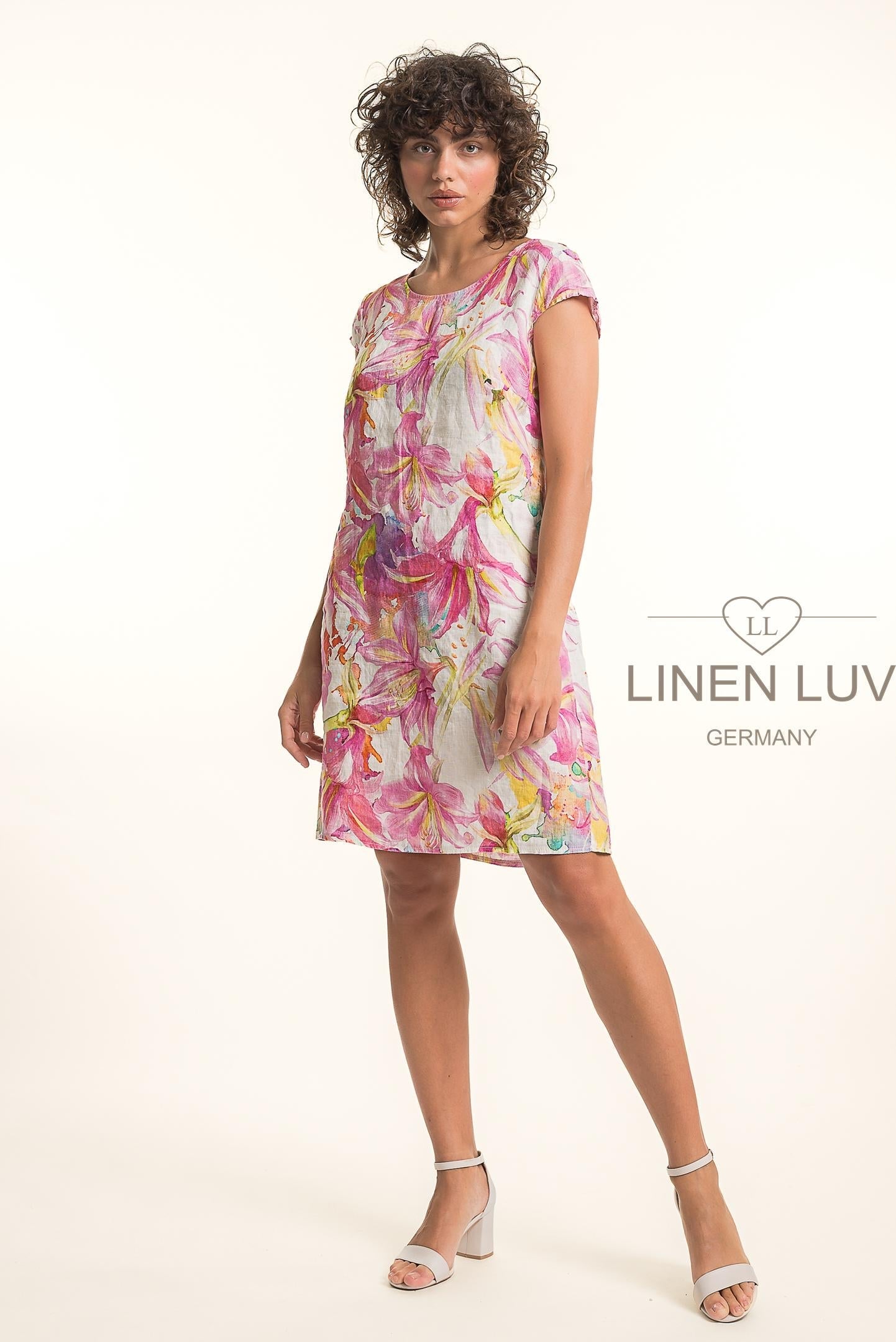 A woman with curly hair stands in a studio wearing the LinenLuv French Linen Dress TP1757P-43 Fuxia, featuring short sleeves and vibrant pink, yellow, and green patterns. She pairs it with white heels against a light background with the LinenLuv logo.