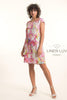 A woman with curly hair stands in a studio wearing the LinenLuv French Linen Dress TP1757P-43 Fuxia, featuring short sleeves and vibrant pink, yellow, and green patterns. She pairs it with white heels against a light background with the LinenLuv logo.