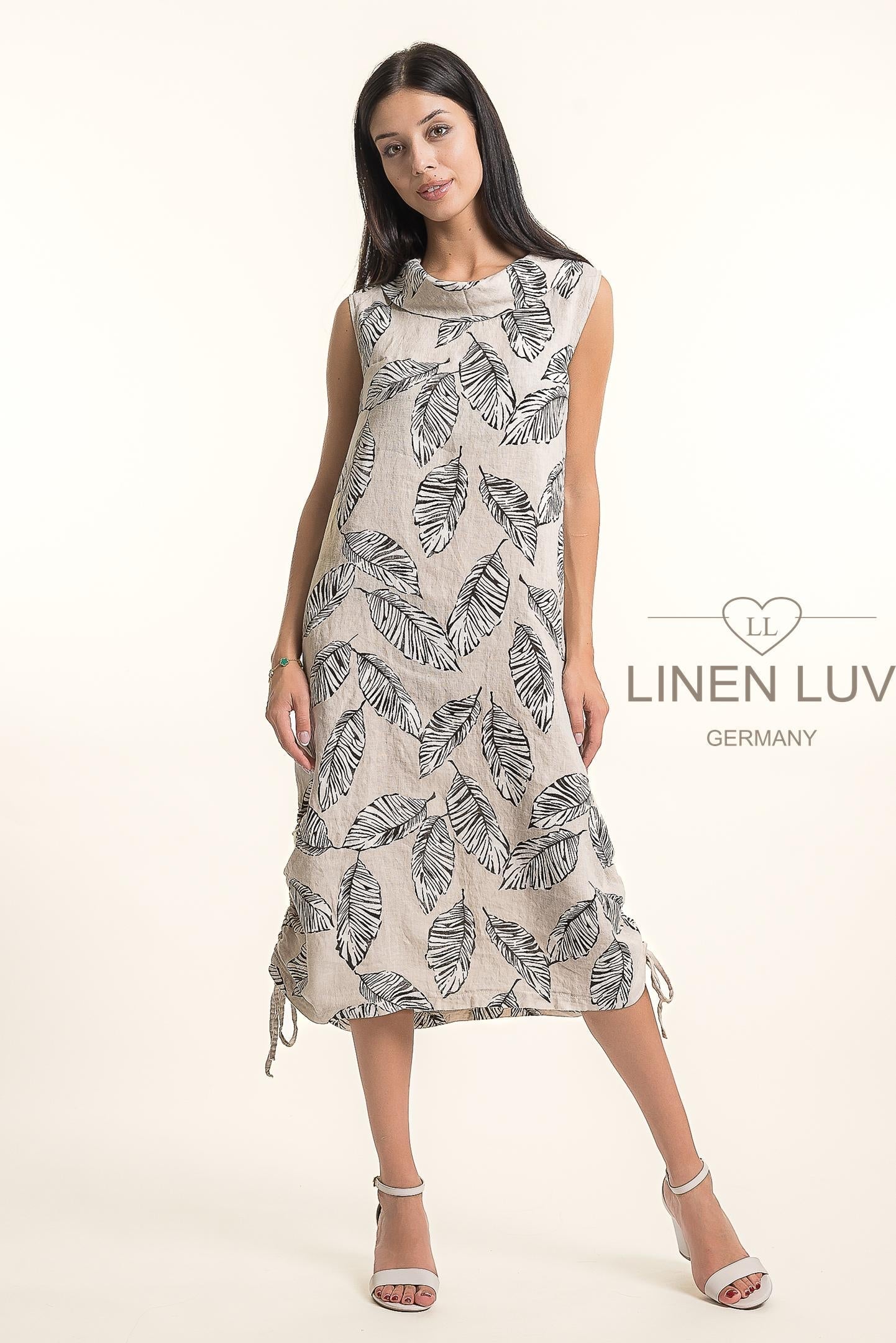 A woman models the French Linen Dress TP1676P-11 Sabbia by LinenLuv, a sleeveless beige dress with a black botanical print, paired with white heels. "LINEN LUV" is displayed to the right.