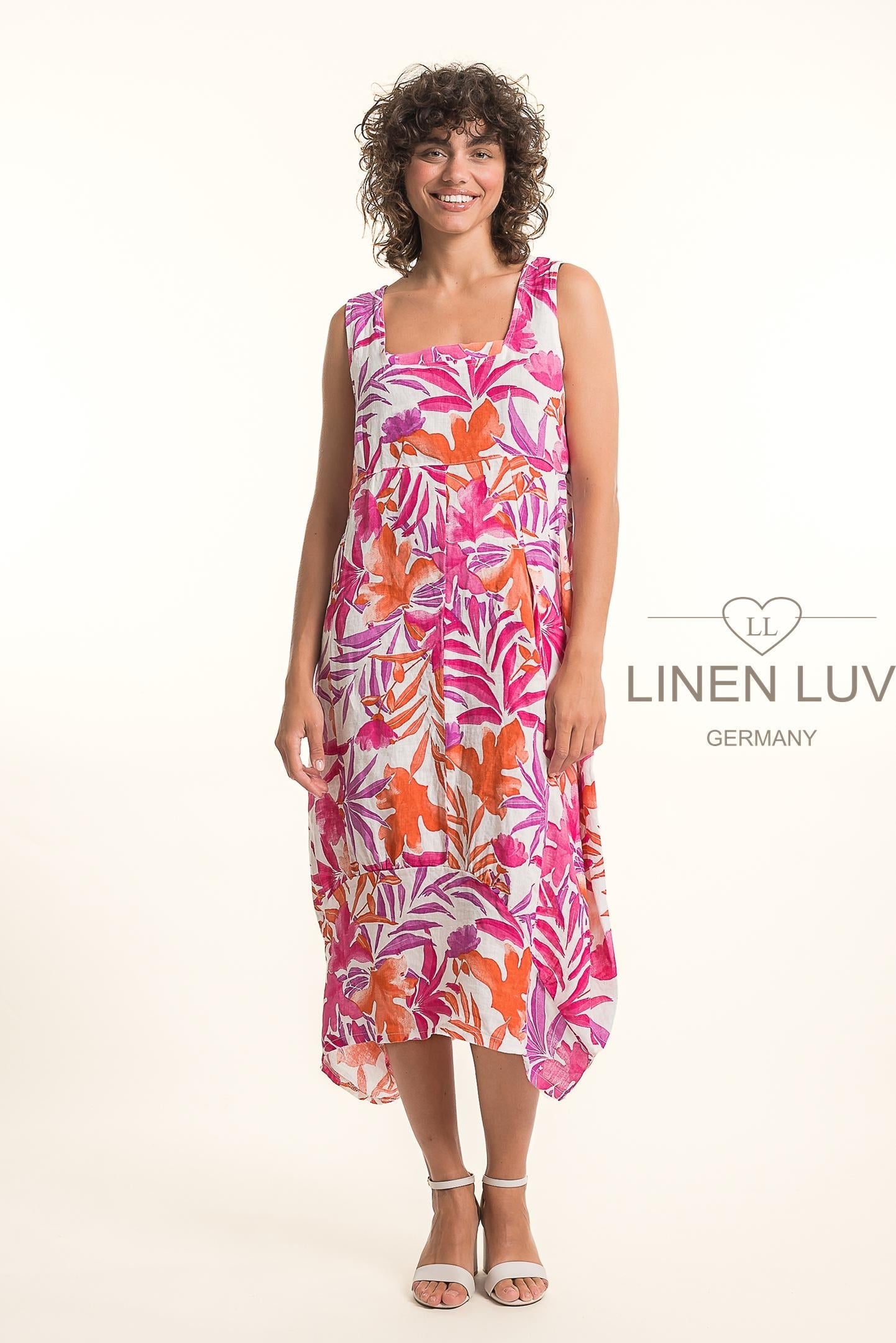 A woman smiles in a sleeveless, relaxed-fit French Linen Dress TP1776P-11 Bianco by LinenLuv, featuring a vibrant pink, orange, and purple abstract floral print. She pairs it with white sandals against a simple white background with the LinenLuv logo.