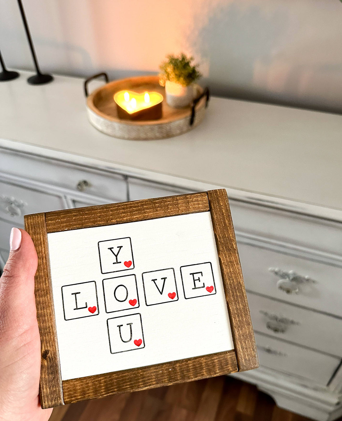 A hand displays the Love You Mini by Inspired Findings, a small wooden sign featuring the phrase “LOVE YOU” arranged crossword-style, with each letter in its own square accented by a red heart. This unique home decor piece is framed in antique walnut and styled alongside a glowing candle and plant on a white dresser.