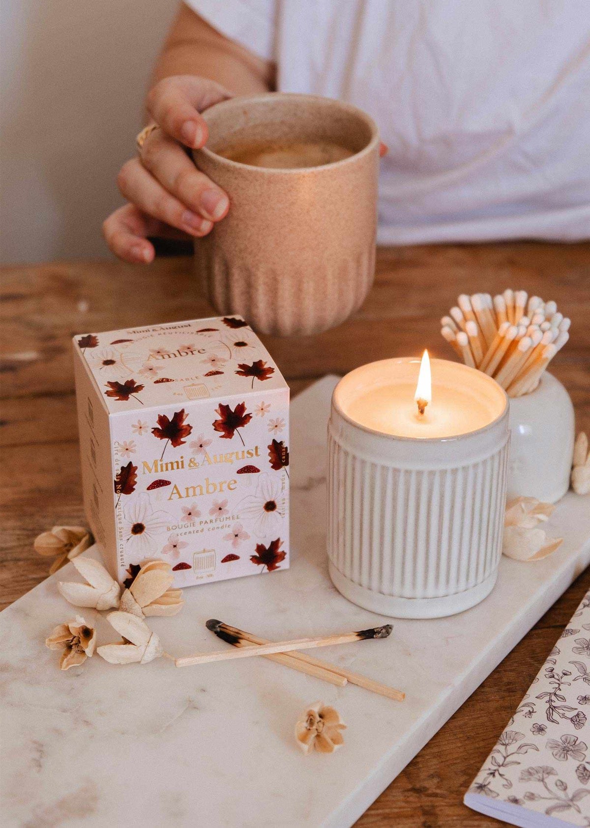A person holding a mug near a lit Mimi & August Ambre - 8 oz Reusable candle, with its box, a jar of matches, and dried flowers arranged on a wooden table.