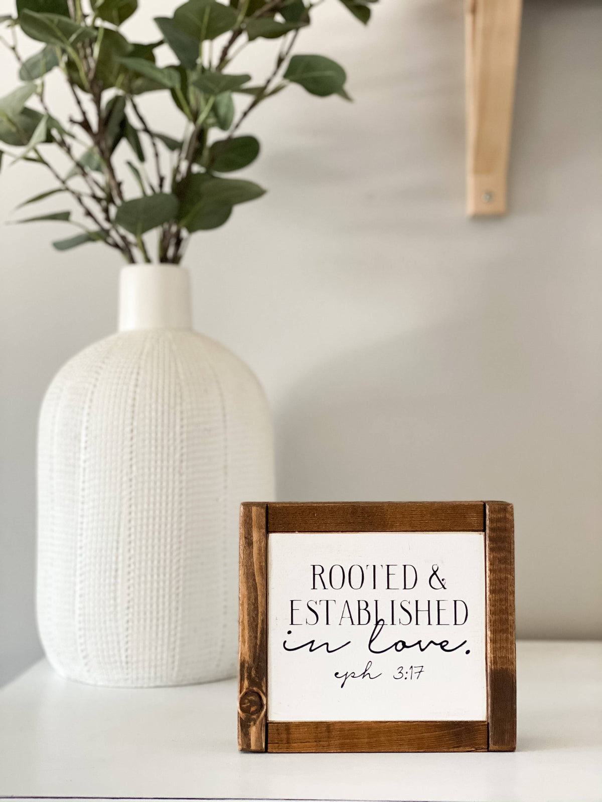 The Inspired Findings “Rooted & Established mini” sign, featuring a wooden antique walnut frame, sits on a white surface with the message “Rooted & Established in love. Eph 3:17.” A textured white vase filled with leafy green branches is placed in the background, complementing the warmth of the wood sign.