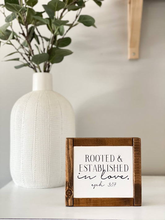 The Inspired Findings “Rooted & Established mini” sign, featuring a wooden antique walnut frame, sits on a white surface with the message “Rooted & Established in love. Eph 3:17.” A textured white vase filled with leafy green branches is placed in the background, complementing the warmth of the wood sign.