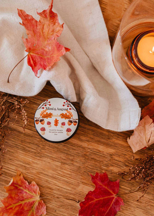 An Ambre - 2 oz Soy Candle by Mimi & August sits on a wooden table draped with a white cloth, accompanied by autumn leaves and a matching tin. The mini amber soy candle fills the space with the inviting warmth and aroma of an autumn forest, creating a cozy fall atmosphere.