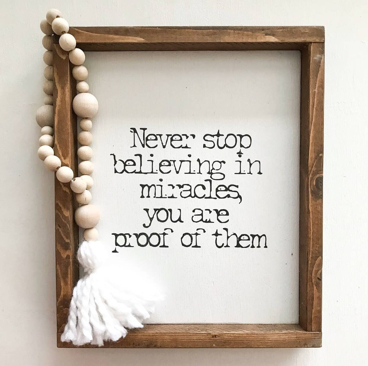 A charming wooden sign from Inspired Findings, featuring a beaded tassel, hangs in the corner. The “Never stop believing in miracles Sm - Inspired Findings” design displays the inspiring words: “Never stop believing in miracles, you are proof of them.”.