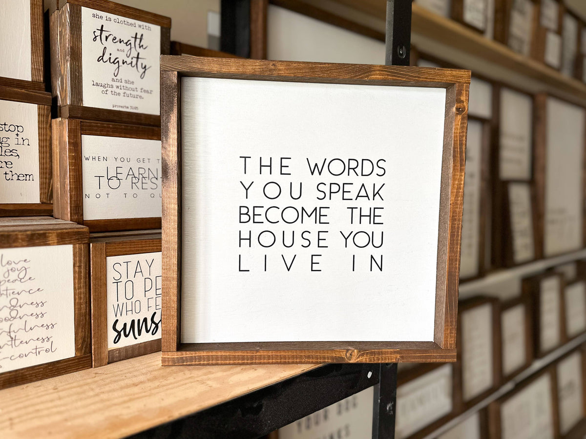 An Inspired Findings wood sign, called “The Words You Speak Wood Sign,” features the phrase “The words you speak become the house you live in,” and is displayed alongside other inspirational decor on shelves.