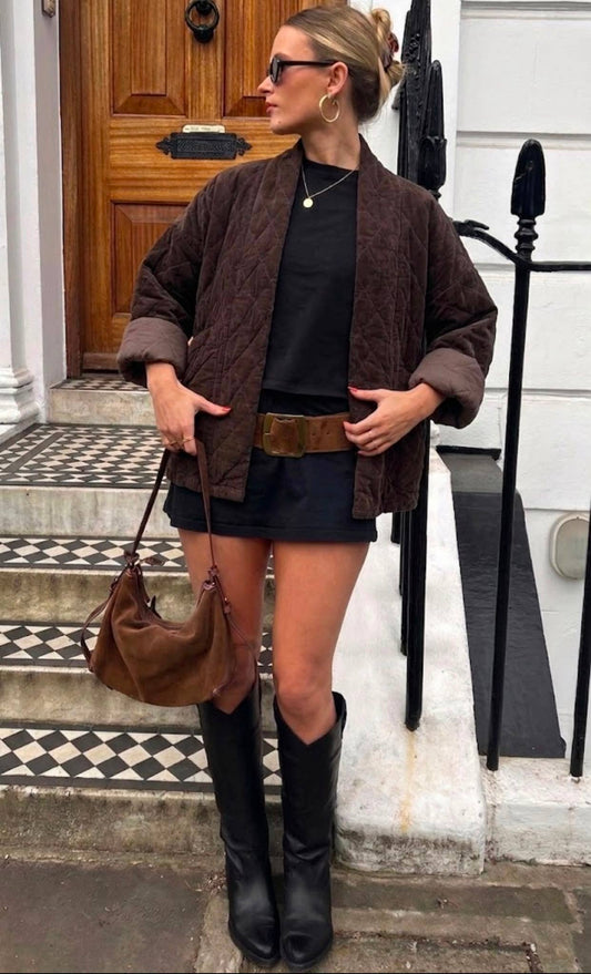 A woman stands on a checkered doorstep in front of a wooden door and white walls, wearing the NOELINE Quilted Jacket 2442-2 in Chocolate. She pairs it with a black shirt, short black skirt, wide brown belt, knee-high black boots, sunglasses, earrings, and carries a brown handbag.