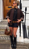 A woman stands on a checkered doorstep in front of a wooden door and white walls, wearing the NOELINE Quilted Jacket 2442-2 in Chocolate. She pairs it with a black shirt, short black skirt, wide brown belt, knee-high black boots, sunglasses, earrings, and carries a brown handbag.