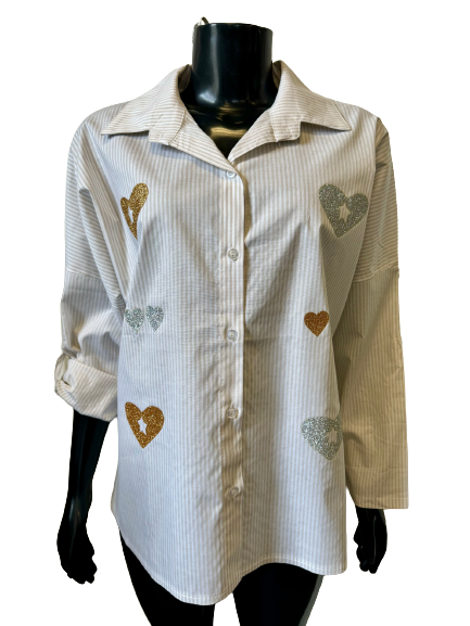 A BUTTON SHIRT - BEIGE STRIPES - ETERNELLE displayed on a mannequin. This long-sleeved, button-up shirt features light gray and white pinstripes adorned with glittery heart-shaped patches in gold and silver on the front. The sleeves are rolled up to three-quarter length.