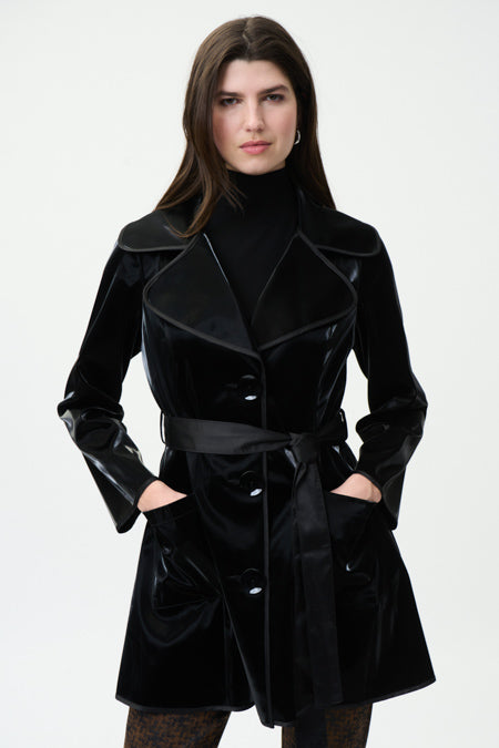 A woman wearing Joseph Ribkoff's Black Synthetic Leather Women's Coat, with a belt tied at the waist, poses against a plain, light background. She has her hands in the coat pockets and is dressed in a black turtleneck underneath. With long, dark hair and eyes fixed on the camera, she epitomizes fall wardrobe elegance.