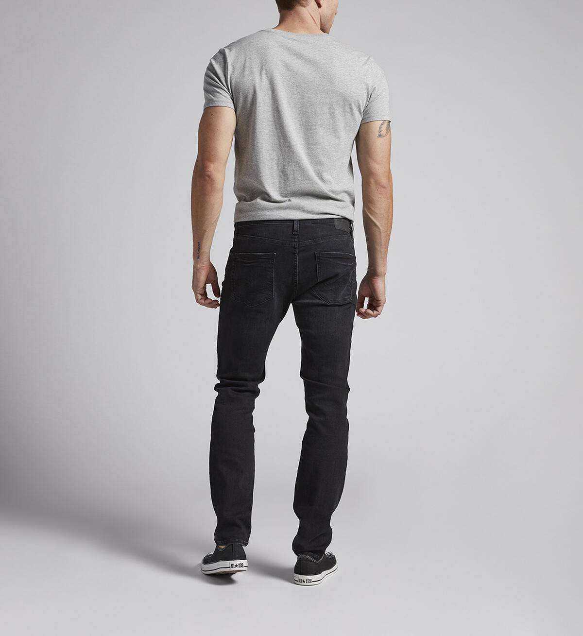 A person with cropped hair is standing and looking to the left in a gray short-sleeve t-shirt, Silver Jeans Co. Taavi Skinny Fit Skinny Leg Jeans in black eco wash made from max flex denim, and black low-top sneakers. The background is plain light gray.