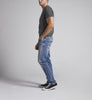 A person standing sideways to the camera showcases the Taavi Skinny Fit Skinny Leg Jeans in medium indigo wash from Silver Jeans Co., paired with a fitted dark gray t-shirt and black and white sneakers against a plain, light-colored background.