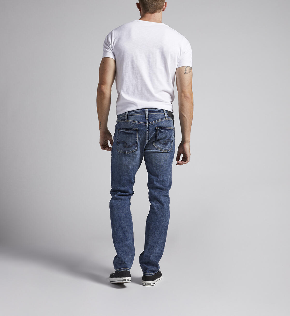 A man is standing against a plain grey background, wearing a white T-shirt, Konrad Slim Fit Slim Leg Jeans from Silver Jeans Co., and black sneakers. His hands are relaxed by his sides, and he is facing slightly to the right. Only the lower part of his face is visible.