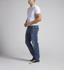 A man stands against a plain grey background. He is wearing a white T-shirt, Konrad Slim Fit Slim Leg Jeans by Silver Jeans Co., and black-and-white sneakers. His hands are clasped in front of him, and he looks slightly to the right.