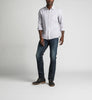 A man stands against a plain background, wearing a light gray button-up shirt with rolled-up sleeves, Konrad Slim Fit Slim Leg Jeans from Silver Jeans Co. in dark indigo rinse, and dark brown shoes. He looks towards the camera with a relaxed posture, with one hand slightly in front.