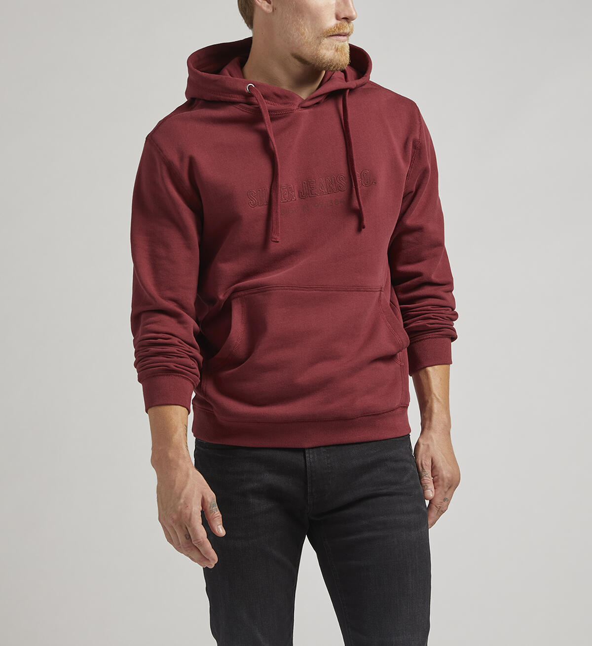 A person with short hair stands facing away from the camera, wearing a Silver Jeans Co. Logo Hoodie in burgundy featuring an adjustable drawstring. They pair it with black jeans and black shoes with white soles. The background is a plain, light gray color.