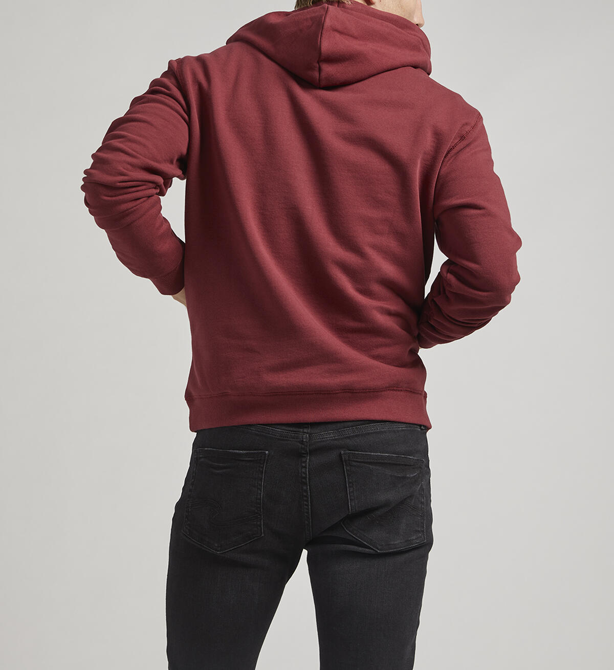 A person with short hair stands facing away from the camera, wearing a Silver Jeans Co. Logo Hoodie in burgundy featuring an adjustable drawstring. They pair it with black jeans and black shoes with white soles. The background is a plain, light gray color.