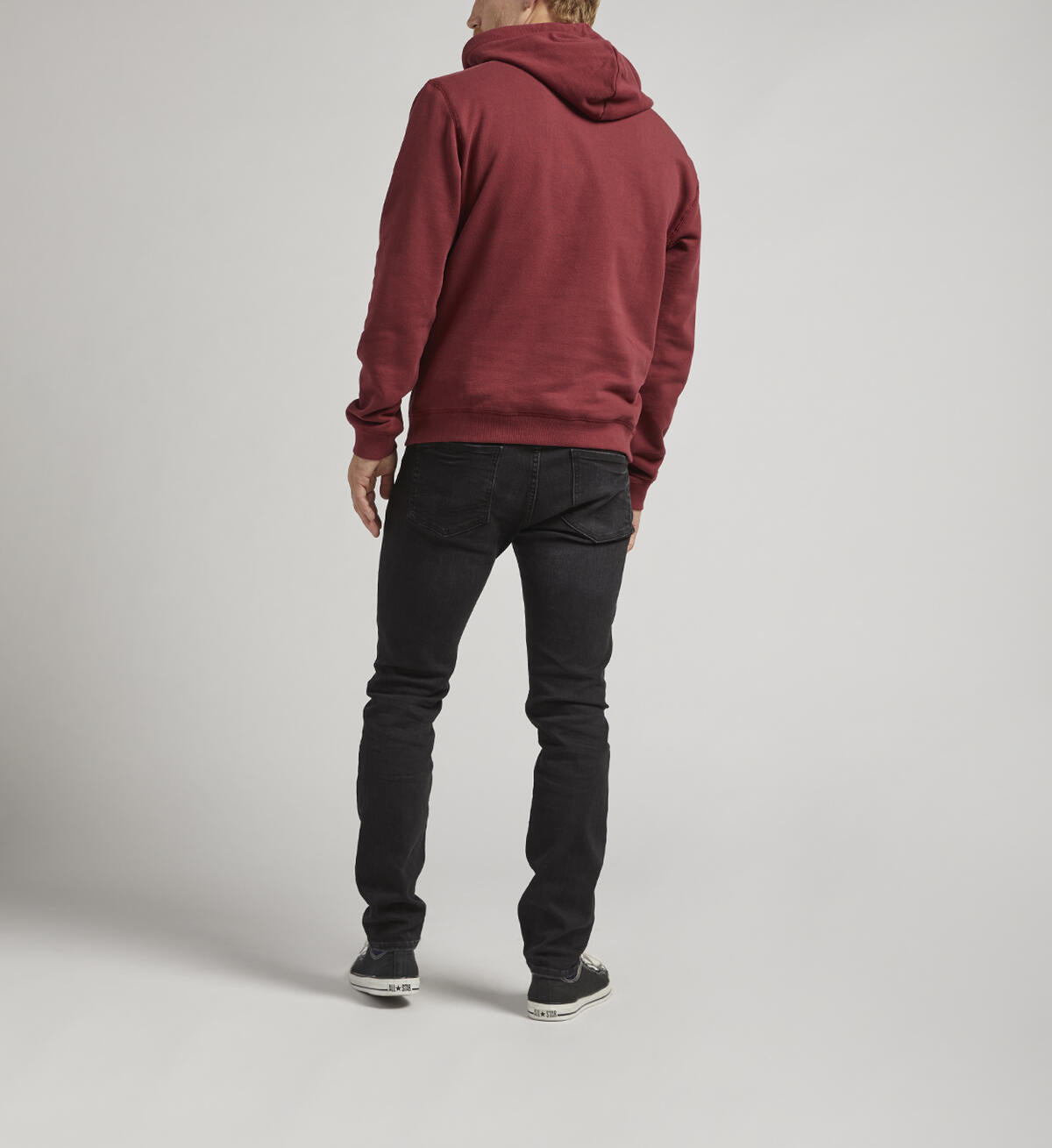 A person with short hair stands facing away from the camera, wearing a Silver Jeans Co. Logo Hoodie in burgundy featuring an adjustable drawstring. They pair it with black jeans and black shoes with white soles. The background is a plain, light gray color.