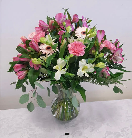 The "Yours Truly" bouquet from Posies Flowers & Fashion features a clear glass vase filled with an assortment of flowers, including pink lilies, white irises, light pink carnations, and small white and pink daisies. This lush arrangement is accented with green foliage and sits elegantly on a white tablecloth. Please note that substitutions may occur based on flower availability.