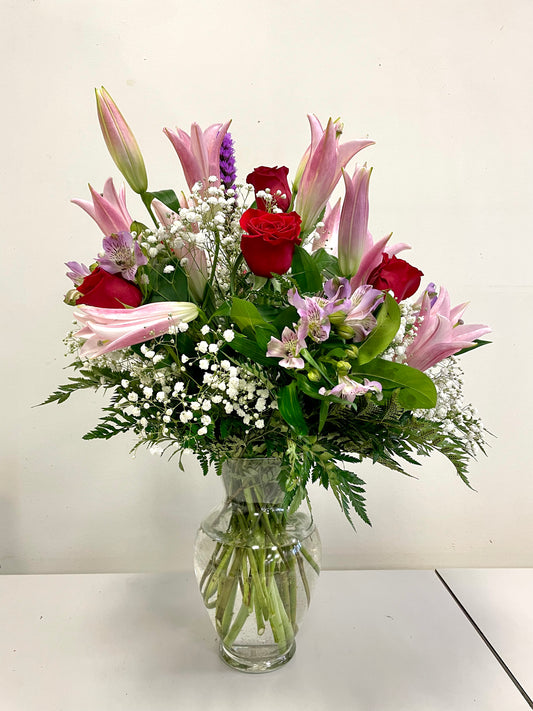 The "Congratulations are in Order" arrangement from Posies Flowers & Fashion showcases a vibrant bouquet in a glass vase, featuring pink lilies, red roses, white baby's breath, and an array of lush green foliage. The flowers are elegantly arranged to create a stunning and fresh display against a plain background. Please note that substitutions may occur depending on seasonal availability.