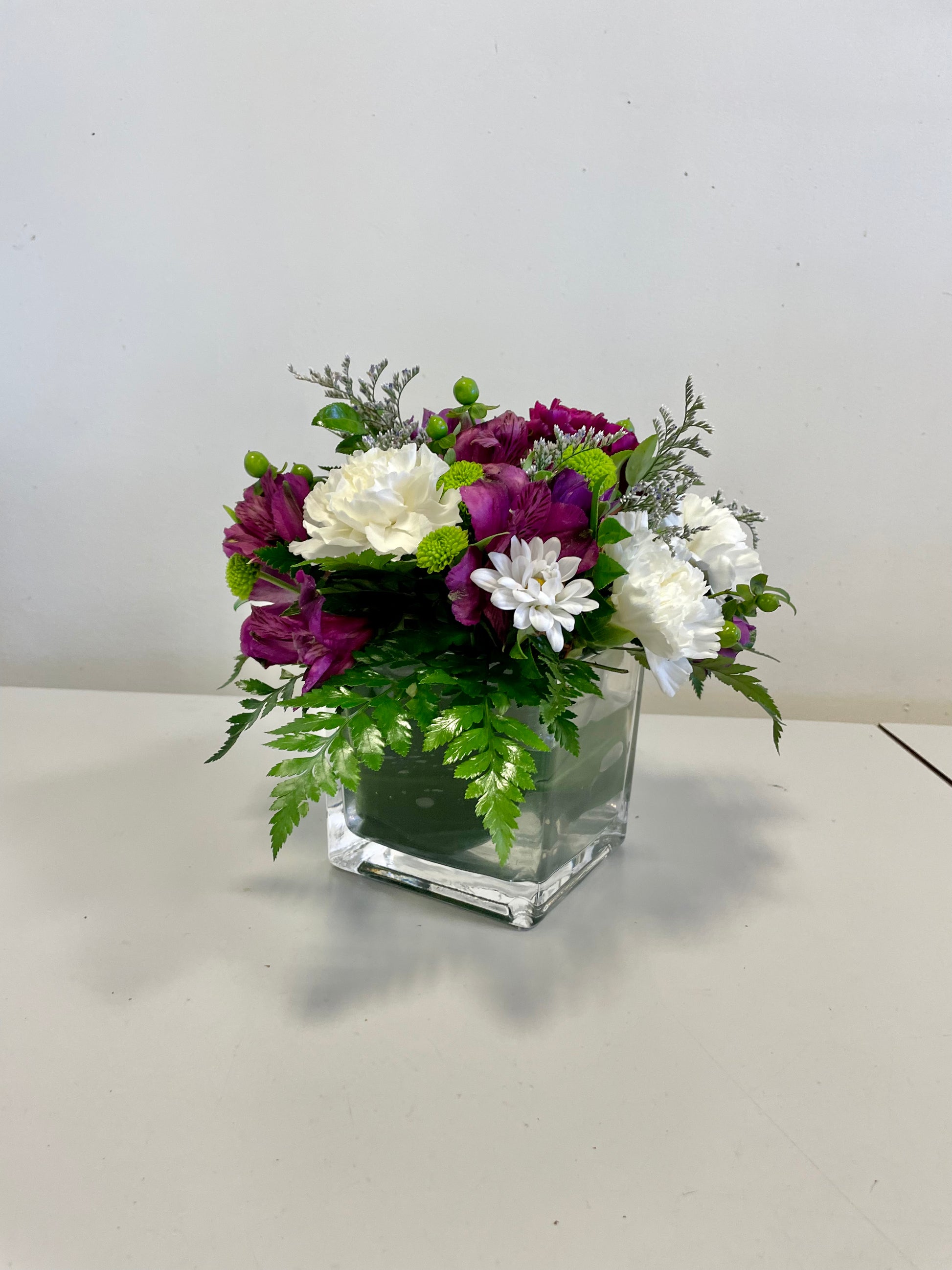 The Beloved Bouquet from Posies Flowers & Fashion is a floral arrangement in a clear square glass vase, featuring a mix of purple, white, and green flowers, including carnations and chrysanthemums, interspersed with green foliage and delicate white fillers. Due to seasonal availability, occasional substitutions may occur. The vase is placed on a white surface against a plain background.