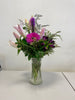 The "Reach for the Sky" bouquet from Posies Flowers & Fashion features a clear glass vase filled with pink lilies, purple daisies, and various types of greenery. Flower substitutions may occur based on availability. The arrangement is set against a plain, light-colored wall backdrop.