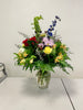 The "Love & Wonder" bouquet from Posies Flowers & Fashion is presented in a clear glass vase, showcasing a vibrant assortment of flowers including red roses, yellow carnations, purple chrysanthemums, and blue delphiniums. Accented with green foliage and ferns for an accurate depiction, this stunning arrangement is displayed on a white table against a plain white wall.
