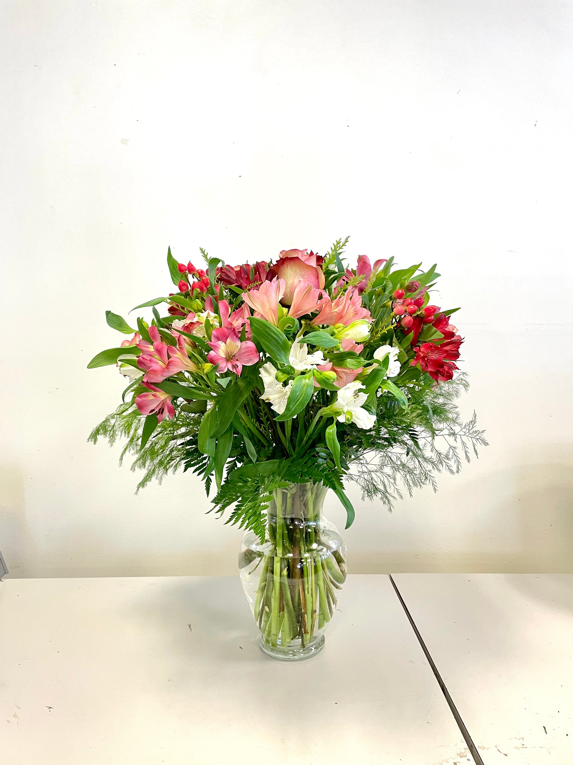 A clear glass vase filled with a vibrant flower arrangement titled "Blushes of Kisses" by Posies Flowers & Fashion. The bouquet features pink roses, red and white lilies, and assorted greenery, all beautifully arranged against a plain white background on a white tabletop.