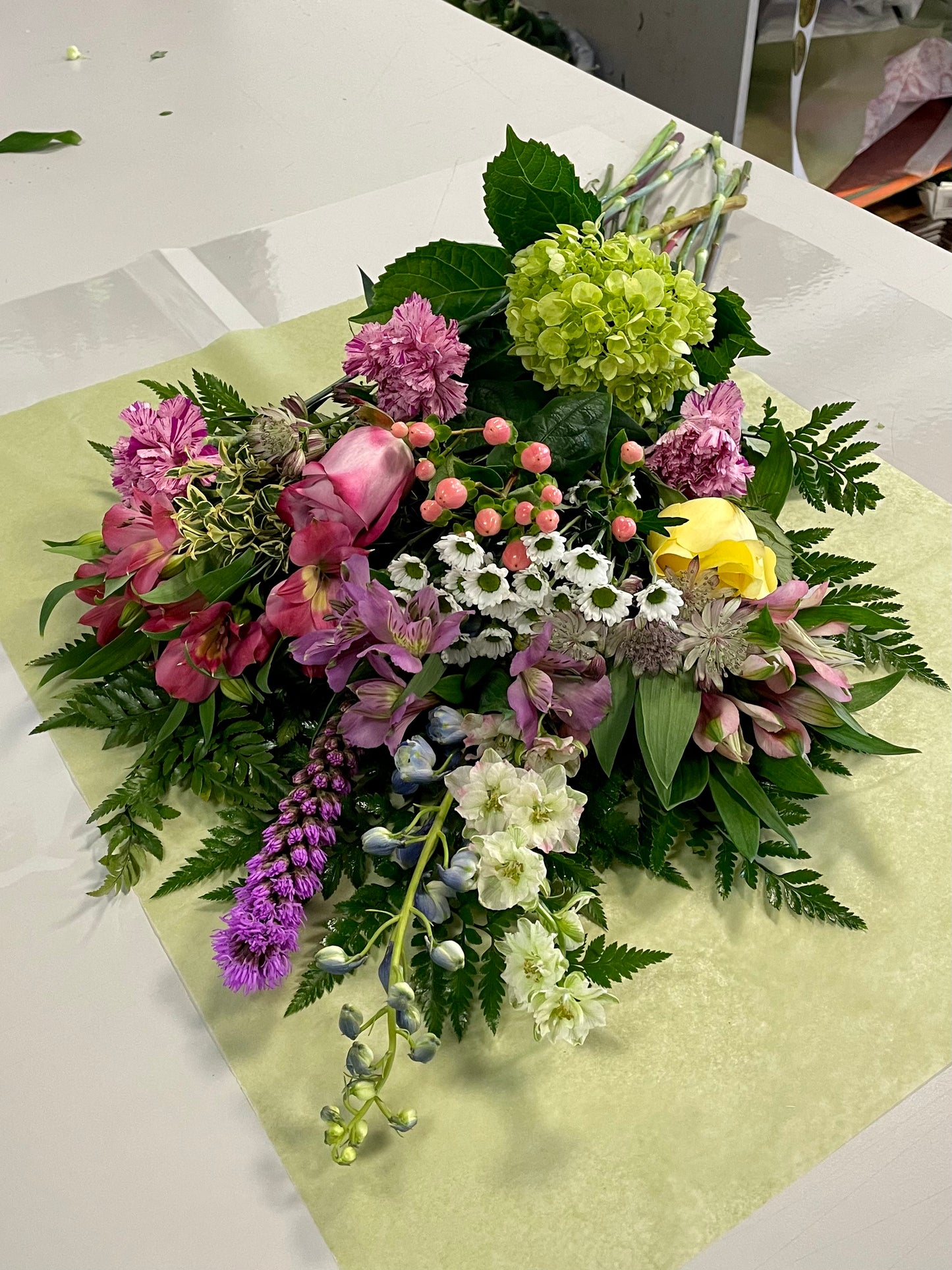 On a table lies a vibrant floral arrangement named "Acknowledge Worthy" by Posies Flowers & Fashion. This stunning bouquet features an exquisite variety of flowers including pink tulips, yellow roses, white daisies, purple liatris, and more, all complemented by lush green foliage. The flowers are beautifully displayed on light green paper, creating an image of pure elegance. Please note that the availability of flowers may vary.