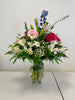 The "Floral Hugs" arrangement by Posies Flowers & Fashion showcases a vibrant mix of pink roses, white lilies, green foliage, and blue delphiniums in a clear glass vase against a plain white background. The flowers and greenery cascade naturally, creating a lush and balanced display; substitutes may be used depending on flower availability.