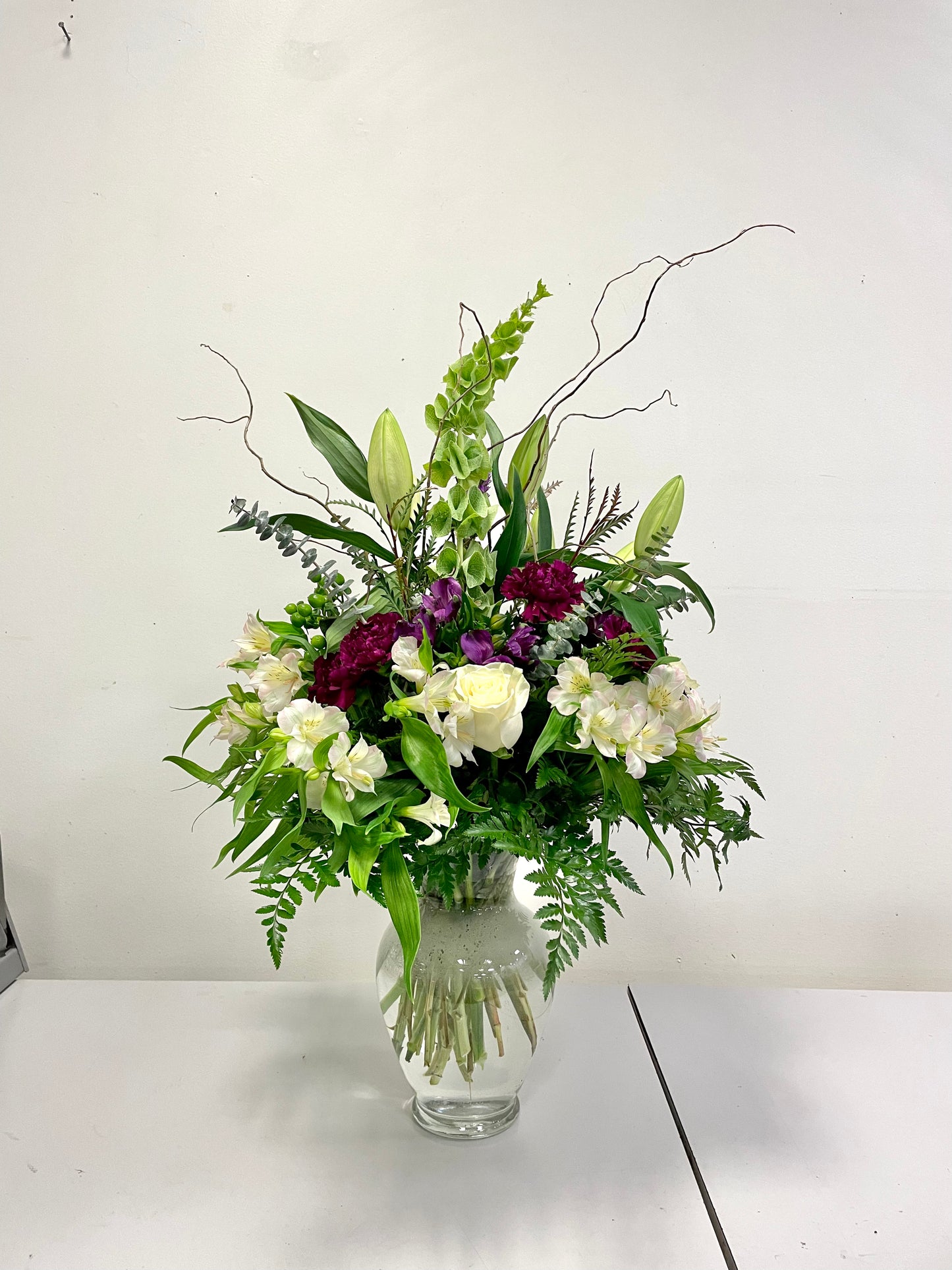 The "In Touch with the Woods" arrangement by Posies Flowers & Fashion features a blend of white, deep purple, and green flowers complemented by a variety of leaves and branches, all elegantly displayed in a clear glass vase. Positioned on a white surface against a plain background, the bouquet's natural beauty shines through without any need for substitutions.