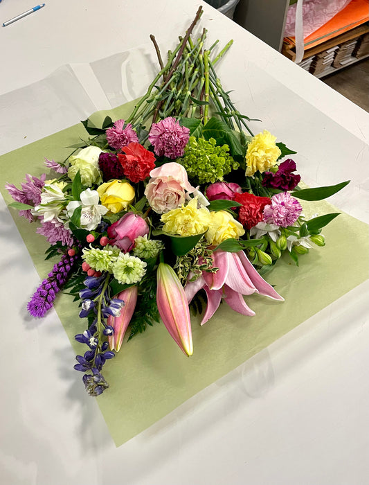 The "Love of Colours Freshcut" bouquet from Posies Flowers & Fashion features pink lilies, yellow roses, red carnations, and purple blooms on light green wrapping paper. Stems are gathered at the top. Substitutions may occur based on flower availability.