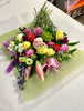 The Love of Colours bouquet by Posies Flowers & Fashion, featuring an array of pink lilies, red and yellow roses, purple and white carnations, and a mix of lush green foliage, is beautifully displayed on a pristine white table. The flowers are artfully arranged with their stems gathered at the top, allowing the blossoms to fan out in a captivating display.