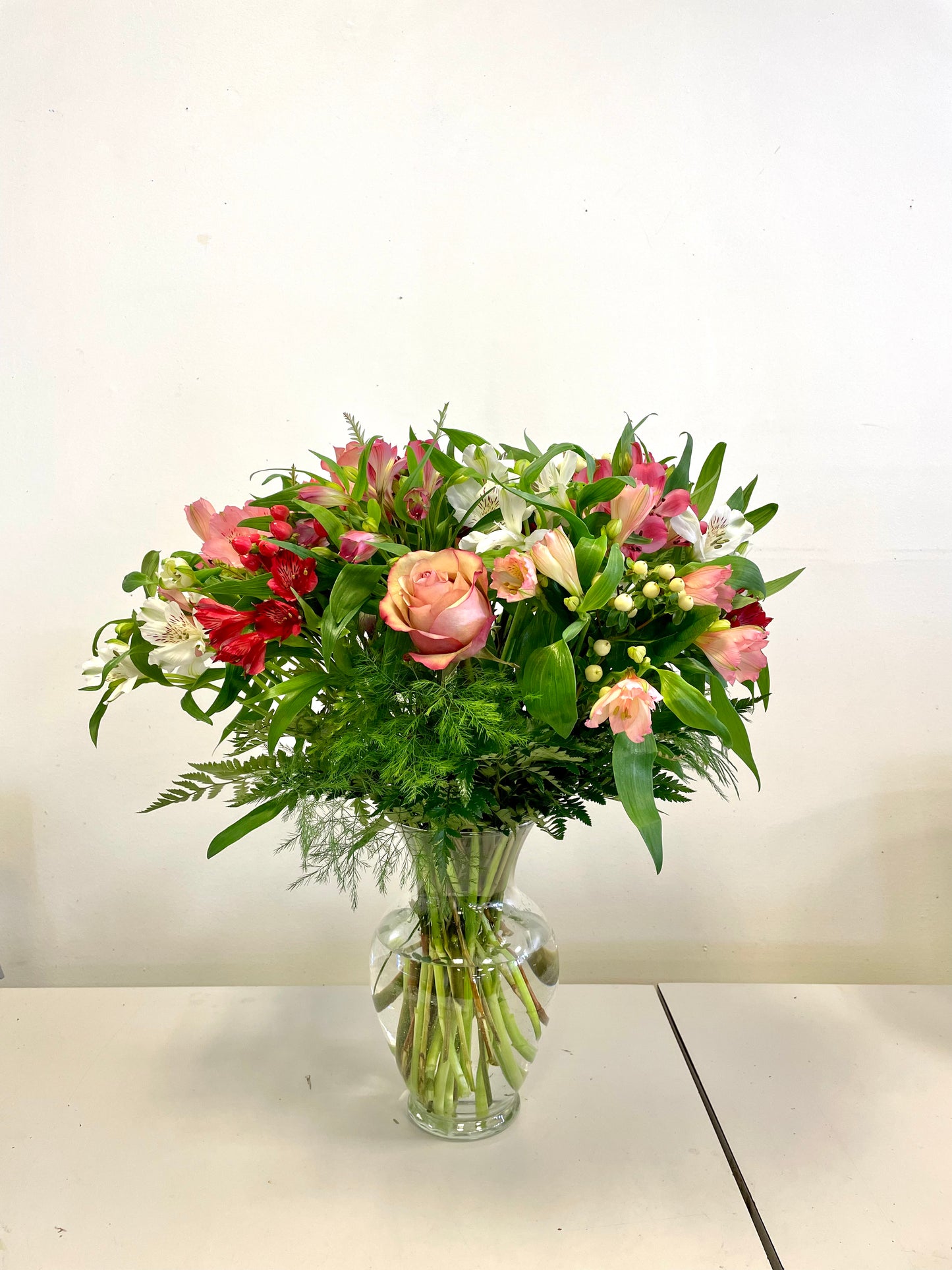 A clear glass vase filled with a vibrant flower arrangement titled "Blushes of Kisses" by Posies Flowers & Fashion. The bouquet features pink roses, red and white lilies, and assorted greenery, all beautifully arranged against a plain white background on a white tabletop.