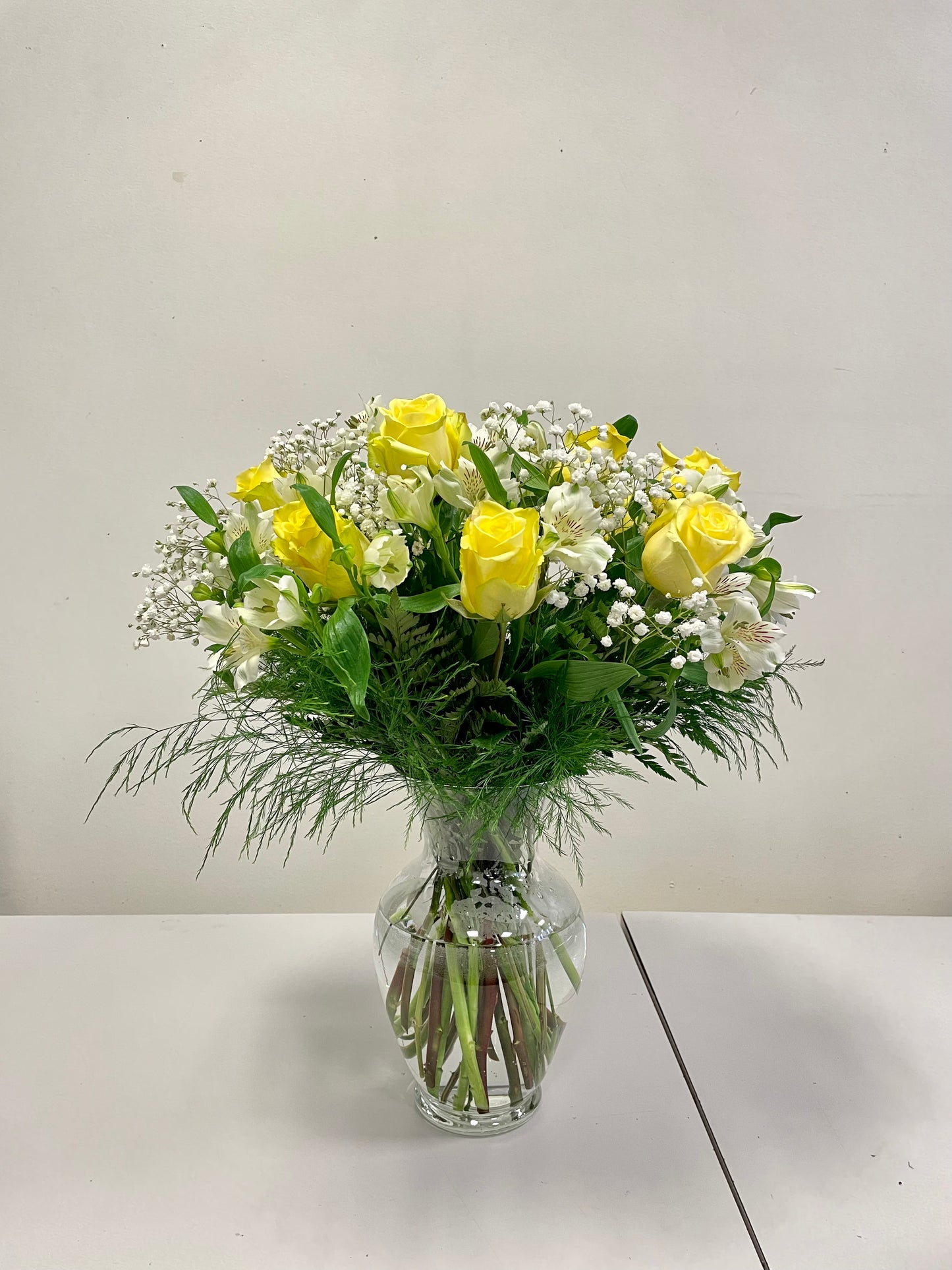 The "Kisses of Sunshine" arrangement from Posies Flowers & Fashion features a glass vase filled with yellow roses, white filler flowers, and greenery. It is displayed on a white tabletop against a plain, light-colored wall. Please note that due to flower availability, substitutions may occur while maintaining the same look and feel.