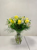 The Kisses of Sunshine Vase by Posies Flowers & Fashion showcases yellow roses, white flowers, and greenery in a clear vase. Substitutions may occur due to availability. The elegant bouquet is beautifully presented against a simple white backdrop on a table for an ideal product image.
