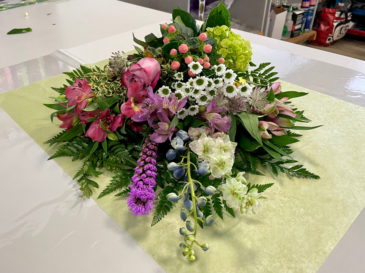 On a table lies a vibrant floral arrangement named "Acknowledge Worthy" by Posies Flowers & Fashion. This stunning bouquet features an exquisite variety of flowers including pink tulips, yellow roses, white daisies, purple liatris, and more, all complemented by lush green foliage. The flowers are beautifully displayed on light green paper, creating an image of pure elegance. Please note that the availability of flowers may vary.